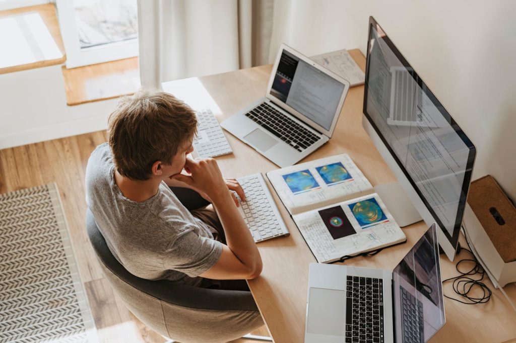 high-angle-shot-of-a-boy-sitting-on-grey-chair-while-using-his-laptop-computers-4974922 Top view of young programmer working on multiple laptops in a modern office setting.
